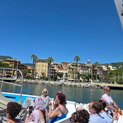 This image depicts a sunny day in Santa Margherita Ligure, Italy, as seen from a boat on the water. In the foreground, several people are seated and standing on the deck of the boat. A woman with dark curly hair and sunglasses is looking to the left. Next to her, a person wearing a light purple headscarf and a floral patterned top is holding a phone and appears to be taking a photo. Another woman with blonde hair and sunglasses is looking forward. Further back, a woman in a pink shirt and sunglasses is holding a colorful scarf. A man in a striped blue shirt is seated. The boat's deck is visible, with railings and ropes.
The background showcases the picturesque harbor of Santa Margherita Ligure. Colorful buildings with red, yellow, and orange facades line the waterfront, interspersed with palm trees. A white statue or monument is visible in the center of the town. The water is calm and reflects the blue sky. Hills covered with green vegetation rise behind the town, and a clear blue sky dominates the upper portion of the image. The scene suggests a leisurely boat tour along the Italian Riviera.
