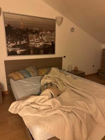 This image depicts an unmade bedroom located in Cava de' Tirreni, Italy. The room features a sloped ceiling indicative of an attic or loft conversion. A large, double-sized bed with a dark wood headboard dominates the center-left. On the bed, white fitted sheets are visible, covered by a light blue flat sheet and two striped pillows (one yellow and blue, one yellow). A beige textured comforter or duvet is heavily rumpled and partially covers the bed, extending towards the foreground.
To the right of the bed, a dark wood nightstand holds a white perforated lamp and a glowing orange Himalayan salt lamp. Above the headboard, a large, rectangular framed print of a nocturnal city view, resembling Paris with the Eiffel Tower illuminated, is mounted on the wall. Two white, upward-facing wall sconces are positioned above and to the right of the framed print. A circular device, possibly a thermostat or smoke detector, is mounted on the sloped white wall to the right of the sconces. The flooring is light-colored wood. In the far right background, a dark wood dresser or chest of drawers is partially visible. No individuals are present in the scene.