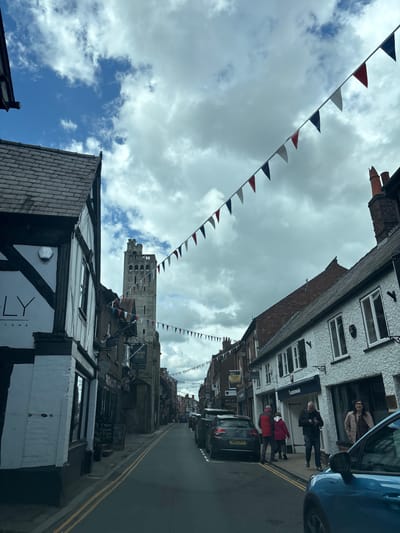 The image shows a street view in Knutsford, United Kingdom. The street is lined with buildings, some of which are Tudor-style with black and white facades. Festive bunting in red, white, and blue hangs across the street. There are cars parked on the side of the road. Several people are walking along the pavement. A tall, stone tower can be seen in the distance at the end of the street. The sky is cloudy, with patches of blue. Double yellow lines are visible in the center of the road.