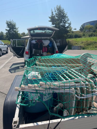 The image captures a white SUV with an attached trailer, both heavily loaded, in an outdoor paved area under a clear blue sky. The scene is located in Sinsheim, Germany, identifiable by the "KLIMA ARENA" building visible in the background, specifically appearing to be near Am alten Auweg 1, 74889 Sinsheim.

The white SUV, possibly a BMW X3, has its rear tailgate and both rear passenger doors open. The cargo area is densely packed with assorted luggage, bags (including a prominent orange backpack), and a dark-colored pet carrier positioned on the left side. No individuals are visible in or around the vehicle.

Attached to the rear of the SUV is a trailer, overloaded with various items and secured by a vibrant green cargo net. Visible contents beneath the net include a large, partially obscured blue spherical object (possibly an exercise ball or large container), multiple white PVC-like pipes or poles, and what appear to be stacked outdoor chairs or furniture components.

The immediate environment consists of a light gray paved surface, likely a parking lot or service road. In the background, beyond a grassy embankment, a large, curved building with "KLIMA ARENA" inscribed on its side is prominent. Tall green trees and power lines are also visible against the bright sky. Shadows indicate a clear, sunny day. Another white vehicle is partially visible on the far left.