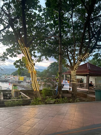 The image depicts a waterfront scene in Bantaeng, Indonesia, at dusk. In the foreground, a tiled walkway leads to the left, bordered by low planters with greenery. The middle ground features several trees, two of which are wrapped with yellow string lights. Behind the trees, a covered pavilion with a reddish-brown roof is visible, with two men seated inside. A body of water lies beyond the pavilion, with a few boats and structures along the shoreline. In the background, hills and mountains are silhouetted against a twilight sky. Several people are scattered throughout the scene, some walking and others seated. The overall atmosphere is relaxed and peaceful, with the lighting suggesting the early evening.
