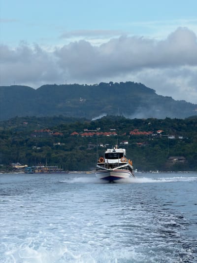 The image shows a boat speeding through the water, creating a large spray. The backdrop features a mountainous landscape with buildings scattered along the hillside. The location is Toyapakeh, Indonesia.