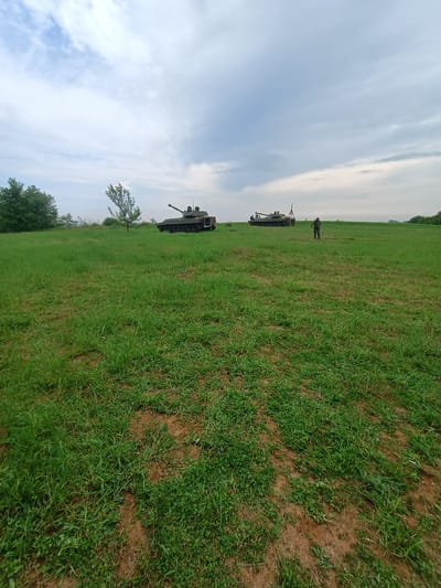 The image shows a grassy field, likely outdoors, with a cloudy sky above. Three tanks are positioned in the distance, spaced out across the field. A single person in military garb stands near the farthest tank. A line of trees is visible in the foreground on the left. The location is possibly Kostena Reka, Bulgaria, due to the request.