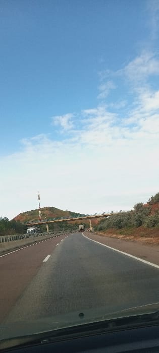 The image displays a multi-lane highway scene in Majmaa Tolba, Morocco, captured from the interior of a moving vehicle. The sky occupies the upper portion, featuring a clear blue expanse with scattered white cumulus clouds. In the immediate foreground, the dark upper edge of a vehicle's dashboard and the lower portion of its windshield are visible. The dark grey asphalt road, marked with white dashed lane lines and a solid white line on the right shoulder, extends centrally into the distance, showing a slight curve to the right. A reddish-brown paved shoulder or median with a white line runs along the left side of the highway, bordered by concrete barriers. In the mid-ground, a beige-colored overpass, supported by visible pillars, spans the highway. Underneath this overpass, a white commercial truck is observed in the left lane, and a darker colored truck or bus is visible in the right lane, both vehicles traveling away from the camera. To the left of the highway, on a reddish-brown, partially vegetated hill, a tall communication tower with red and white segments stands prominently. The environment surrounding the highway consists of reddish-brown hills with sparse, low-lying green vegetation. No pedestrians or clearly discernible individuals are visible. The scene indicates active traffic flow on a rural or semi-rural motorway.