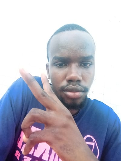 A close-up portrait captured in Mtopanga ward, Kenya, features a single male subject. The individual has dark skin, short dark hair, and a trimmed beard and mustache. He is wearing a blue short-sleeved t-shirt with a partially visible, faint pink/purple graphic design on the left side of his torso. The subject's left hand is raised, with the index and middle fingers extended upwards in a 'V' gesture, while the ring and pinky fingers are curled. His thumb is also visible, partially obscuring the lower left portion of his face. The subject is looking directly towards the camera. The background is uniformly bright white, appearing to be a plain wall, with subtle horizontal and vertical lines suggesting a tiled or textured surface faintly discernible in the upper and lower right portions of the frame.