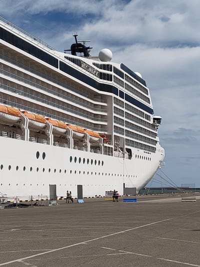 The image shows the side of the cruise ship "MSC Musica" docked in the port of Cagliari, Italy, with a view of the sky with some clouds. The ship's white hull features rows of round porthole windows and orange-covered lifeboats. People are walking on the dock between the ship and the parking lot. The dock is made of asphalt with painted parking lines. The water of the harbor is visible behind the ship.
