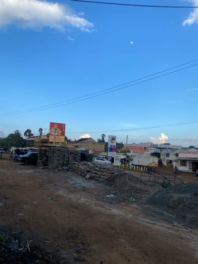The scene depicts a daytime street in Kianyaga, Kenya, under a clear blue sky featuring scattered white clouds and a visible crescent moon. Overhead power lines traverse the upper portion of the frame.
The foreground consists of an unpaved, dusty ground surface. Multiple stacks of grey concrete blocks and several piles of loose gravel are present. A black metal frame structure lies in the right foreground.
In the midground, to the left, a large billboard displays a Christmas-themed advertisement featuring a figure in red. Below the billboard, a dark-colored sedan is parked on the dirt. Several individuals are discernible throughout the midground, some near vehicles. To the right, a Riva petrol station is prominent, with its sign displaying fuel prices: 198.80, 192.50, and 187.90. A white truck and another light-colored truck are positioned near the station's pumps, along with a white passenger vehicle. Adjacent to the petrol station, a white building labeled "GOLD GLASSMART DUKA LTD" is visible, with a person on a motorcycle in its vicinity.
The background shows green tree lines and scattered buildings on gently rolling terrain.