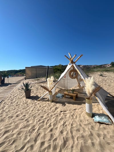 The image depicts a tranquil beach scene in Dyuni, Bulgaria, under a clear blue sky. In the foreground, a white teepee-like tent is set up on the sand, adorned with decorative elements like a dreamcatcher and dried pampas grass in a golden vase. A low wooden table sits inside the tent, holding a small plant and what appears to be a tray with items. Cushions are scattered around the tent, one light blue cushion resting on the sand near the vase.
To the left of the tent, a wooden walkway leads towards the sea, with a large potted agave plant situated next to it. Beyond the walkway, a sheltered structure, possibly a beach bar or restaurant, is visible. In the distance, the beach is lined with rows of sun loungers and colorful umbrellas, suggesting a resort area. The background features a gentle slope covered in green vegetation and buildings on the hillside, overlooking the Black Sea. The sand is textured with footprints and small shells, indicating recent activity. The overall impression is one of a peaceful, well-decorated space prepared for relaxation on a sunny day.