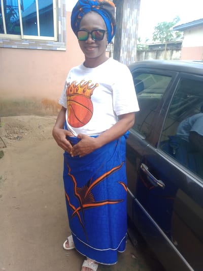A woman stands outdoors in Obehie, Nigeria, facing the camera with a warm smile. She is wearing a vibrant blue, orange, and black patterned headwrap, complemented by mirrored sunglasses. Her attire includes a white t-shirt featuring a graphic of a basketball adorned with a golden crown, and a matching blue, orange, and black patterned wrapper skirt. White, open-toed sandals are visible on her feet, which rest on sandy, unpaved ground. Her hands are clasped together in front of her midsection, and a ring is visible on her left ring finger.
The setting appears to be a residential area during daylight hours, under bright, sunny conditions. Behind the woman, to her left, is a light-colored building with a blue-tinted window. To her right, a dark-colored car is parked, partially visible, reflecting the surroundings and a figure in a blue shirt in its window. In the background, beyond the car, there's a fence, some trees, and other buildings, suggesting a neighborhood environment. The hem of the wrapper skirt shows faint text that reads "VERITABLE PRINTED BY".