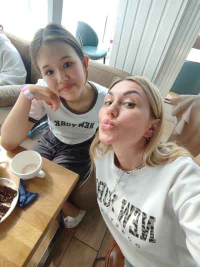 Two females are photographed from the chest up, in a selfie style, indoors, likely in a cafe or restaurant. The female in the foreground, on the right, has blonde hair and is making a kissing gesture towards the camera. She is wearing a white sweatshirt with a leopard print design on the letters that spell out "NEW YORK". She also has a silver necklace and earrings. The female in the background, on the left, has brown hair pulled back and is wearing a white t-shirt with "NEW YORK" printed on it. She has a pink and white wristband on her left wrist. They are seated at a wooden table. On the table, there is a white coffee cup with some liquid in it, a blue napkin, and a plate with a slice of chocolate cake. A water bottle is visible behind the girl on the left. The background shows a couch, a chair, and vertical wooden paneling. The image quality is moderate, with some blurring due to the selfie pose. There is no indication of the location being Amursk, Russia.