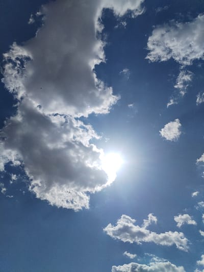 This media file captures a dynamic view of a bright blue sky filled with a variety of clouds. The sun is prominently visible, partially obscured by a large, fluffy white and gray cloud formation on the left side of the frame. Sun rays emanate outwards, creating a lens flare effect.
Scattered throughout the sky are smaller, wispy white clouds, some appearing more dense and cumulus-like, while others are thin and spread out. A tiny dark silhouette of what appears to be a bird is visible in the upper left quadrant, adding a sense of scale and life to the vast sky.
The lighting suggests it is daytime, likely midday or afternoon, given the intensity of the sun. The weather appears to be fair with some cloud cover, typical for a sunny day. No specific location cues are visible beyond the expanse of the sky. The overall mood is bright and open, conveying a sense of a pleasant day. There is no visible text in the image.