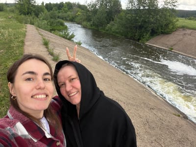 Two light-skinned women are positioned close together, smiling at the camera. The woman on the left wears a black hooded sweatshirt and holds up two fingers in a peace sign. The woman on the right wears a plaid shirt over a white top. They are standing on a concrete edge next to flowing water. Below them is a wooded area. The location is Nizhnii Vishur, Russia.