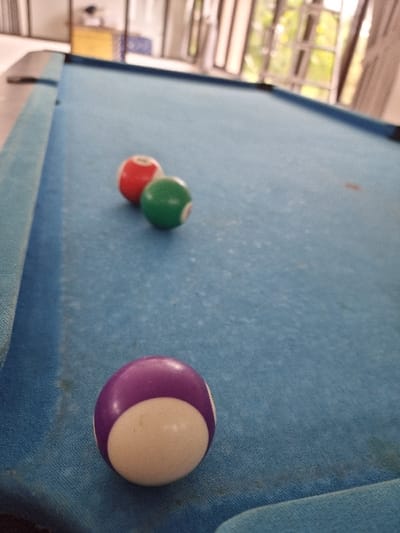 The image displays a blue-felted pool table located indoors in Sindangjaya, Indonesia. Three pool balls are present on the worn surface of the table. A purple and white striped ball is positioned prominently in the foreground, on the lower left side. Further up the table, a solid red ball and a solid green ball are situated near each other, slightly left of the table's longitudinal center. The blue felt exhibits visible dust and light discoloration. The background is out of focus, revealing a large window structure with natural light and hints of outdoor greenery, along with blurred light-colored objects, including one yellow item, in the far distance. No individuals are visible, and no active play is depicted.