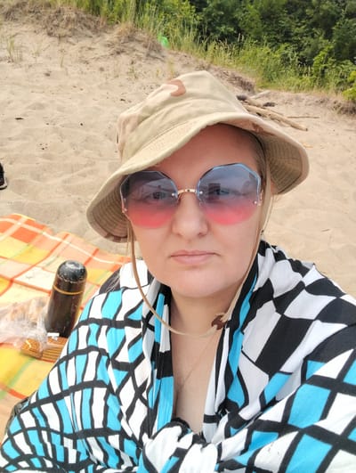 A woman is sitting on a beach in Bibikha, Russia, with a picnic blanket spread out on the sand. She is wearing a camouflage bucket hat and oversized, octagonal sunglasses with a pink and blue gradient. A black and white patterned scarf is draped around her shoulders. To her left, there is a dark thermos and a plastic bag containing crackers. The background shows sand dunes and lush green trees.