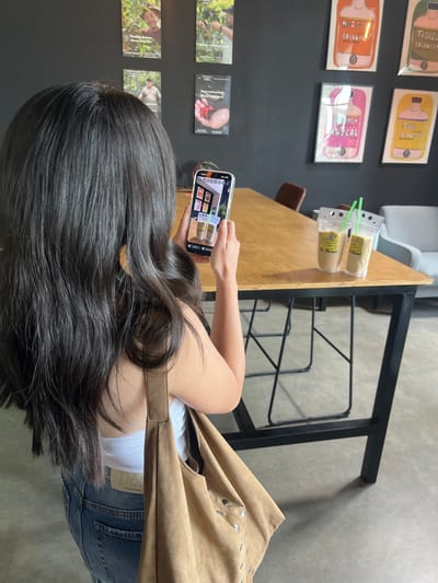 A person, viewed from behind, with long dark hair, wearing a white tank top, blue jeans with a "D&G" label, and carrying a tan suede-like shoulder bag, is holding a silver smartphone horizontally to photograph two clear beverage pouches on a wooden table. The phone screen displays the image being captured. Each pouch contains a light brown liquid and a green plastic straw. A yellow label on one pouch reads, "We Brew & Sell Coffee on-site at El Paso." The background wall, dark gray, features framed artwork: four posters on the left depict coffee cultivation, and four prints on the right illustrate stylized alcohol bottles labeled "Whiskey Coldbrew," "Tequila Coldbrew," "Mezcal," and "Rum Coldbrew." A brown chair is partially visible behind the table, and a light gray armchair is in the distant background. The scene is an interior of a cafe or coffee shop in El Paso, United States.