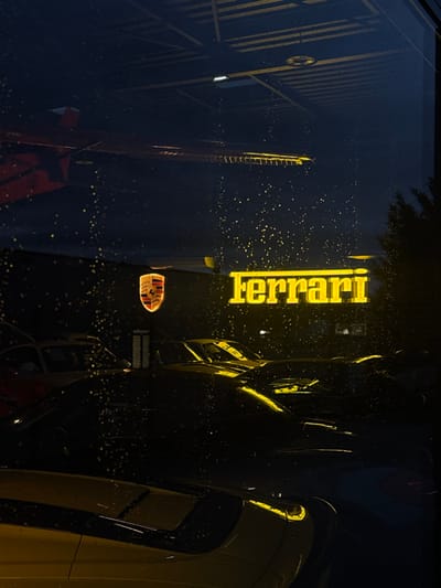 The image displays an exterior perspective, at night, looking into a car dealership showroom in Souffelweyersheim, France, through a glass window. The window surface is covered with numerous small water droplets, creating a diffused, bokeh effect across the view.
Inside the darkly lit showroom, two illuminated corporate signs are prominently featured. A horizontal, yellow neon sign spelling "Ferrari" in its characteristic font is positioned centrally. To its left and slightly above, an illuminated, shield-shaped emblem bearing the Porsche crest is visible.
Below these signs, multiple high-performance or luxury vehicles are parked. In the immediate foreground, the front section of a yellow sports car, including parts of its hood and fender, is visible, reflecting some light. Further within the showroom, additional vehicles, appearing as dark shapes with glints of reflected light on their surfaces, are indistinctly arranged; one appears to be a classic yellow sports car further back.
Reflections are present in the upper section of the glass, including a red angular object on the upper left and a horizontal structure with multiple segments above the Ferrari sign, possibly reflecting internal showroom architecture. Outside the showroom, a dark mass of foliage, likely a tree or dense shrubbery, is visible on the far right. No individuals are present in the scene.