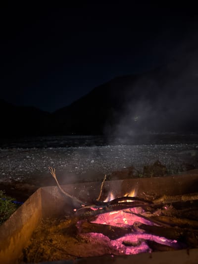The scene is a nighttime view in Sindieti, Georgia. A body of dark water is visible on the left side of the image. A concrete embankment separates the water from a contained fire burning within a metal structure. The fire emits smoke. The fire's flames are partially obscured by tree branches.