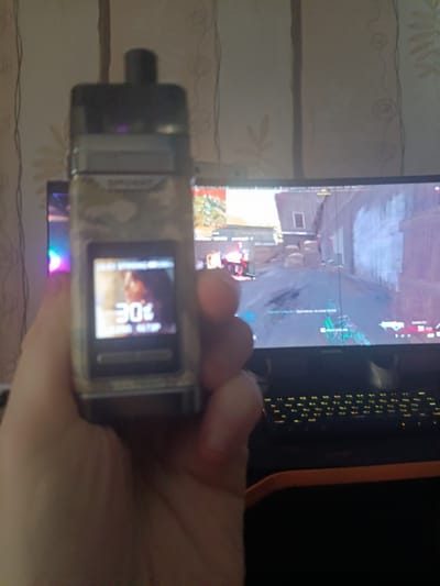 The image shows a close-up of a hand holding a vape device with a digital screen displaying the number "30". In the background, a computer monitor displays a video game scene, and a keyboard is visible below the monitor. The environment appears to be indoors, with a patterned wallpaper visible on the wall. The location of Шипуново, Russia is not discernible from the image itself.