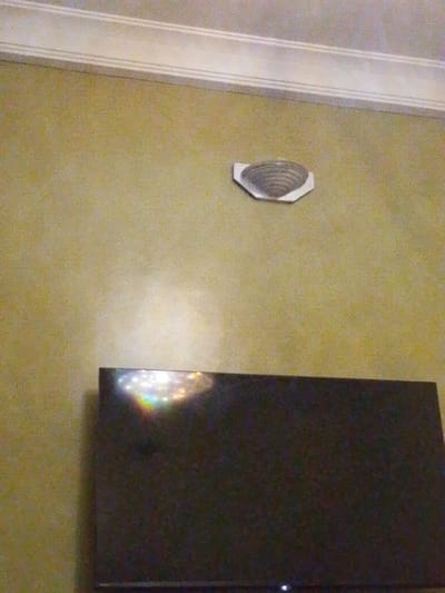The image depicts an interior wall in a room, likely a living area. The wall has a yellowish-green textured paint finish. Along the top of the wall, there is a white decorative crown molding with two horizontal indentations.
Mounted on the wall, slightly above and to the left of center, is a hexagonal white fixture with a metallic, concentric-ringed insert. This appears to be a smoke detector or a similar ceiling-mounted sensor.
Below the fixture and towards the bottom of the image, a dark, flat-screen television is visible. The top edge of the television reflects light, showing a rainbow-like spectrum, suggesting the presence of lights or a light source above and in front of the TV. The television is not on, but a small white indicator light is visible at its bottom edge.
The scene is devoid of any people or discernable action. The environment suggests a domestic setting. The location of Rumuomasi, Nigeria, is not explicitly indicated by any visual cues in the image itself.