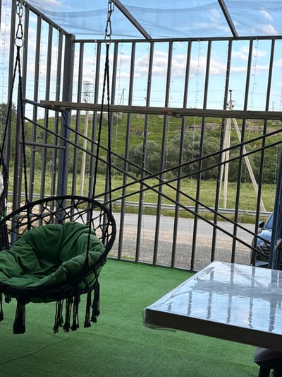 This image depicts an outdoor seating area, likely on a balcony or patio, in the city of Tereze, Russia. In the foreground, a square table is covered with a reflective, clear plastic sheeting. To the left, a green-cushioned, black macrame hanging chair is suspended from the ceiling, with its tassels reaching down to the artificial green turf flooring. Behind the chair, a metal fence structure creates a barrier. Beyond the fence, a lush green hillside is visible, dotted with trees and wildflowers. A road runs along the base of the hill, and a dark-colored car is partially visible to the right. Overhead, a clear plastic roof is supported by a metal frame, and strings of small, white lights are attached to the structure. The sky above is a pale blue with scattered white clouds. There are no people visible in the image.