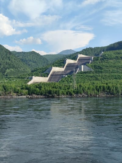 The media file shows a wide, dark river in the foreground, with its surface rippled by a gentle current. In the middle ground, a massive concrete dam complex is built into the side of a steep, tree-covered hill. The dam has a complex, stepped design with multiple levels and sloped surfaces. A tall metal electricity pylon stands in front of a section of the dam, suggesting the presence of a power station. The dam is surrounded by lush green trees, indicating a natural and somewhat remote setting.
In the background, more tree-covered hills rise, with a distant, hazy mountain peak visible on the far left. The sky is a bright blue with scattered white clouds, suggesting it is daytime and the weather is fair. The overall scene depicts a large-scale hydroelectric dam integrated into a mountainous, forested landscape. There is no visible text in the media file. The setting appears to be Cheremushki, Russia, known for its hydroelectric power facilities.