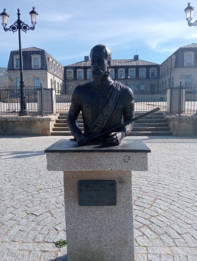 A bronze bust of Fernando Álvarez de Toledo y Pimentel, the 3rd Duke of Alba, is centrally positioned on a light gray speckled granite pedestal in a public square in Piedrahíta, Spain. The bust depicts a male figure with a beard and mustache, attired in a decorated doublet and a diagonal sash. His hands rest on a dark rectangular surface atop the pedestal, with his right hand holding a rod or rolled document. A dark rectangular plaque on the pedestal's front identifies the subject and indicates an installation date of October 2019. The ground is paved with light gray cobblestones arranged in an arched pattern, with sparse weeds visible. In the background, a large stone building, consistent with the Palacio de los Duques de Alba, features multiple windows and a dark mansard roof with dormers. This building is separated from the foreground by a black metal fence and stone steps. Two ornate black lampposts with multiple lanterns are visible on either side of the statue. The scene is brightly lit by direct sunlight, casting distinct shadows.
