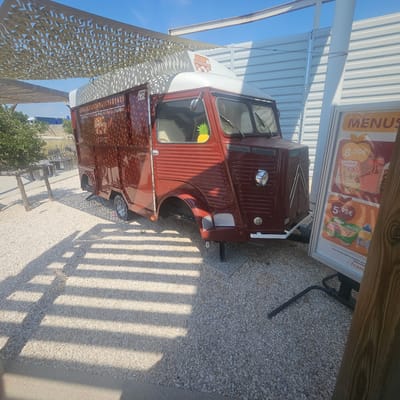 An exterior, low-angle shot captures a retro, maroon-colored food truck parked on a light-colored gravel lot. The truck features a white roof and has a graphic of a different food truck on its side, along with text that reads "LE SPOT." A pineapple sticker is visible inside the driver's side window. To the right of the food truck, a stand displays a menu board with various items and prices, including "8,50€" and "5,95€." The scene is partially shaded by a light-colored canopy with a patterned design, casting striped shadows across the ground. In the background, a corrugated metal wall and some greenery are visible, suggesting an outdoor establishment. The location is identified as La Palme, France.