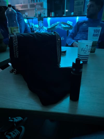 A table illuminated by blue light displays several items. A vape pen lies next to a white cup featuring a rooster design. A large black bag with silver chain detailing dominates the scene, partially obscuring a water bottle. A person in a blue shirt is visible in the background. A pair of black and white sneakers rests on the floor near the table.