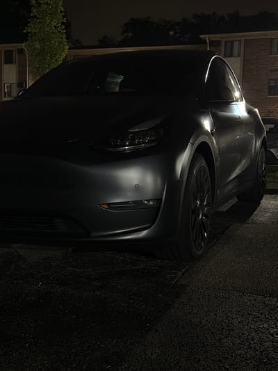The image shows a dark-colored Tesla parked at night in what appears to be a residential area, possibly an apartment complex in Vernon Hills, United States of America. The car is the primary subject, with its front and side visible. The lighting is dim, with some artificial light sources reflecting off the car's surface, highlighting its curves and the headlights. In the background, there are parts of buildings and trees. The ground is asphalt, possibly a parking lot or a street. There are no people or any other vehicles clearly visible in the frame.