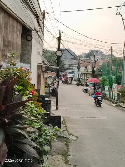 The image depicts a street scene in Bojong Gede, Indonesia. In the foreground, lush green plants and dark red foliage are visible on the left side of the frame, bordering a gray paved walkway and a stone wall. The street itself is asphalt, with some cracks and unevenness.
Further down the street, several people are present, mostly on motorcycles. A person in a red shirt is riding a motorcycle towards the camera. To the left of the street, there are food carts and other vendors. One cart has a large red umbrella. People are seen interacting with vendors and riding their motorcycles. Buildings line both sides of the street, some with a more modern design and others appearing older. Overhead, a dense network of electrical wires is strung between poles, a common sight in many urban and suburban areas. The sky is overcast with a soft, diffused light, suggesting it might be early morning or late afternoon. A timestamp at the bottom left of the image reads "2026/06/21 11:10".