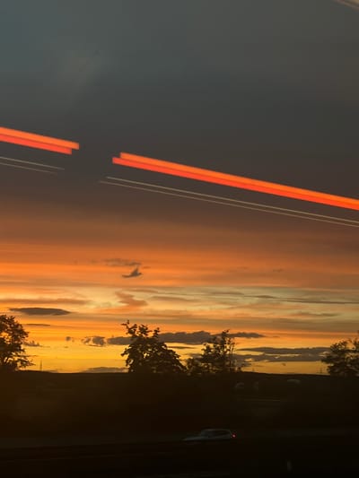 This is a vertical, nighttime photograph taken from a moving vehicle, likely on a highway or road near Bubenreuth, Germany, as indicated by the presence of light trails from other vehicles. The sky is dominated by a dramatic sunset, with hues of deep orange and yellow bleeding into a dark, twilight blue. Wispy clouds are scattered across the sky, catching the last rays of sunlight. In the foreground, a dark silhouette of trees and landscape is visible, with a faint outline of a vehicle on the road below. The light trails, appearing as streaks of orange and white, suggest the motion of traffic. There are no discernible people or specific landmarks that identify the exact location within Bubenreuth.
