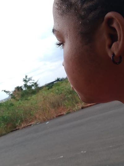 The image presents a close-up, side-profile view of a person with dark skin and black, braided hair. The person is wearing a small, black hoop earring in their right ear. The individual's eye is visible, showing dark eyelashes. In the background, there is a paved road and green vegetation, including tall grasses and some trees. The sky is overcast with light-colored clouds. The location is OGBOKO, Nigeria, judging from information provided.