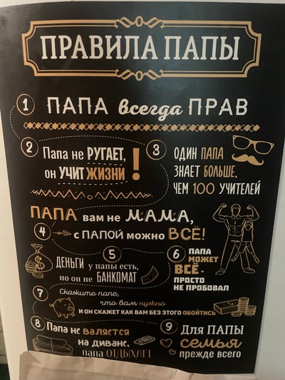 This is a close-up shot of a black poster with gold and white lettering, featuring a list of "Rules of Dad" in Russian. The poster is likely decorative and intended for a family setting.

The main subjects are the text and accompanying illustrations. The text is presented in a playful and humorous tone, with numbered points outlining supposed dad-related wisdom. For example, point 1 states "Dad is always right," and point 8 says "Dad doesn't lie on the couch, Dad relaxes." There are also small illustrations, such as glasses and a mustache, a muscular male figure, stacks of coins, and a piggy bank, which visually complement the text.

The scene depicts a wall, likely in a home, with the poster as the central focus. There are no obvious cues about the time of day or weather. The overall emotion conveyed by the poster is lighthearted and affectionate towards fathers. The text is clear and legible, highlighting the humorous "rules" of being a dad. The location context "Сарженка, Russia" suggests the poster might be found in a Russian household.