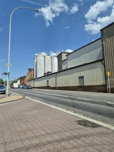 This is a daytime, eye-level, outdoor shot of a street scene in Käerjeng, Luxembourg, likely near a commercial or industrial area. A long, grey corrugated metal building dominates the right side of the frame. To the left of this building are several large, cylindrical storage silos, also metallic and white. In the foreground, a paved street runs horizontally across the image, with a brick-patterned sidewalk on the left. Several cars are visible on the street, moving away from the camera. A white van with the word "FIOR" on its side is also present. A lamppost stands tall on the sidewalk, its curved arm extending over the street. A traffic signal with a red light is visible further down the street. The sky is blue with scattered white clouds. The overall impression is of an ordinary day in a functional urban or industrial landscape.