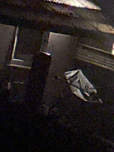 A grainy, dimly lit overhead image, identified as being taken in Galway, Ireland, depicts an urban environment, possibly an alleyway or courtyard, under low light conditions. The scene is dominated by dark tones with structural elements and objects partially illuminated or reflective.
**Structural Elements:**
To the upper right, a light-colored, textured edge, consistent with a roofline or awning, is visible. In the mid-left, a distinct L-shaped light-toned frame is present against a dark vertical surface, potentially outlining a door or window. Adjacent and slightly to the right of this frame, a rectangular opening with a lighter interior suggests a doorway or passage. Towards the mid-right, a dark, linear structure, possibly a bench, steps, or railing, is partially seen against a dark wall.
**Objects:**
Near the rectangular opening in the mid-center, a small, elongated object with a bright, possibly blue-white glowing tip is discernible. In the lower right quadrant, a crumpled, light-gray or off-white fabric or plastic sheet, resembling a tarp or blanket, rests on the dark ground. Faint, indistinct light reflections or small items are minimally visible in the lower-left.
**People/Actions/Interactions:**
No human figures, animals, or vehicles are present. There are no discernible actions or interactions occurring within the frame.
**Environment:**
The image quality indicates very low ambient light, suggesting nighttime or a deeply shadowed area. The surfaces appear consistent with urban materials such as pavement and building facades. The exact location within Galway, Ireland, beyond the general city context provided, is not ascertainable from the visual evidence.