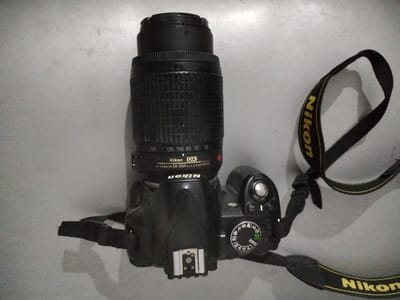 The media file is a top-down, slightly angled, high-resolution photograph of a black Nikon DSLR camera with a telephoto zoom lens attached. The camera is resting on a plain, textured grey surface, likely a table or countertop. A black camera strap with yellow "Nikon" lettering is draped across the camera and the surface.

The lens is a Nikon DX AF-S NIKKOR 55-200mm 1:4-5.6G ED VR. Focal length markings are visible on the lens barrel, ranging from 55mm to 200mm. The camera body shows a typical DSLR layout with a mode dial on the top, featuring various shooting modes such as Auto, Guide, Manual, Aperture Priority, and Shutter Priority. Buttons for "Off" and "On," along with an "Info" button, are also visible. A hot shoe for an external flash is present on top of the camera body. The camera's serial number or model indicator appears to be visible on the side, reading "D3100".

The lighting is flat, suggesting an indoor setting without strong directional light sources. The shadows are soft, indicating the light is diffused. There are no people in the frame, and the focus is solely on the camera and its accessories. The overall impression is that of a product photograph, perhaps for sale or review, with no specific environmental context beyond the grey surface. The photograph was taken in Moara Domnească, Romania.