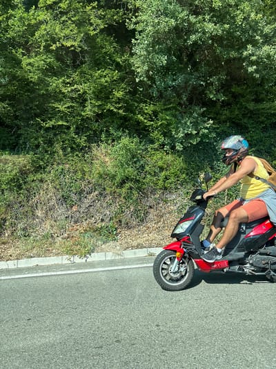 A person wearing a yellow t-shirt, grey shorts, white socks, and black sneakers is riding a red and black scooter. They are also wearing a helmet with a blue, white, and black design. The person is leaning forward, holding onto the handlebars. The scooter is on an asphalt road with a curb on the left side. To the left of the road is a dense area of green trees and bushes. The image is taken outdoors during the daytime, as indicated by the bright sunlight and shadows. The weather appears to be sunny and warm. The scene suggests a casual ride through a natural, possibly rural, or semi-rural environment.