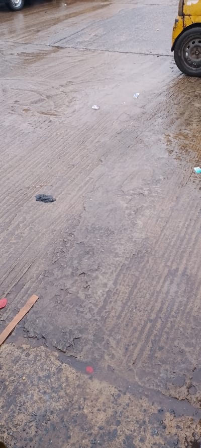 The image depicts a wet, textured asphalt surface, likely a road or parking lot, with visible tire tracks and some peeling sections. A yellow vehicle is partially visible on the right side, its wheel and part of its body suggesting it is a truck or utility vehicle. Scattered across the surface are small pieces of debris, including a crumpled dark object, a thin brown strip, and a small red circular object partially submerged in a puddle. The overall impression is of an outdoor, possibly urban, environment after rainfall, given the presence of puddles and the wet sheen on the ground. The lighting and the wet conditions suggest it might be daytime, possibly overcast. The scene is devoid of people and focuses entirely on the ground and the partial view of the vehicle, creating a somewhat desolate or utilitarian atmosphere. The location context provided places this scene in Jibowu, Nigeria, which might imply the type of vehicle and road surface commonly found in that area. There is no visible text in the image.
