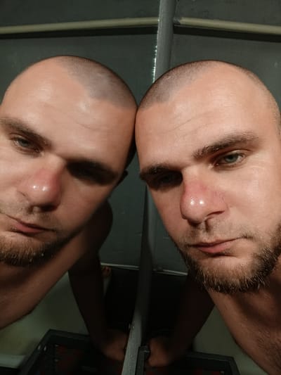 The image is a close-up, slightly angled, selfie-style photograph of a man's head and upper torso, taken in what appears to be a dimly lit indoor environment. The man has a shaved head and a short beard. His face is partially illuminated, highlighting his eyes, nose, and mouth. The man's right eye is visible, with a greenish-blue iris. His nose is slightly red, and there are visible pores and some shine on his skin, possibly due to sweat or oil. His lips are closed.

The image is a mirror reflection, showing two views of the man side-by-side. The background is a dark, textured wall with a horizontal pipe running across the top. Below the man's reflection, a dark object, possibly a piece of furniture or equipment, is visible.

The overall impression is a candid, unposed shot. The location is not discernible from the image itself, and the mention of Pishchane, Ukraine, is not supported by any visual cues within the photograph.