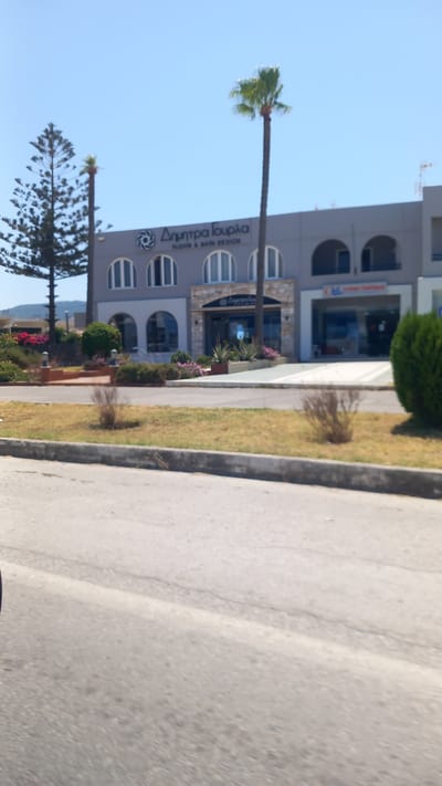 A sunlit, slightly angled, medium shot captures a commercial building with a modern facade situated in Μαρμαρωτό, Greece. The building is painted a light grey, with a series of arched windows lining the upper floor. A large sign with Greek lettering, "ΔΗΜΗΤΡΑ ΓΟΥΡΛΑ," and the English text "FLOOR & BATH DESIGN" is prominently displayed above the central entrance. To the right of this entrance, another shop is visible with the text "CHL" and "HOLIDAYS" on its signage, suggesting a travel agency. The building's entrance is adorned with stone accents and planters filled with greenery and pink flowers.
In front of the building, a manicured lawn separates it from a paved road. A tall, slender palm tree stands to the right of the building, and a more mature, fuller tree with a dark trunk and feathery foliage is positioned to the left. Additional landscaping, including bushes and flowering plants, enhances the area. The sky above is a clear, vibrant blue, indicating a bright, sunny day. The overall impression is one of a well-maintained commercial establishment in a coastal or Mediterranean setting.