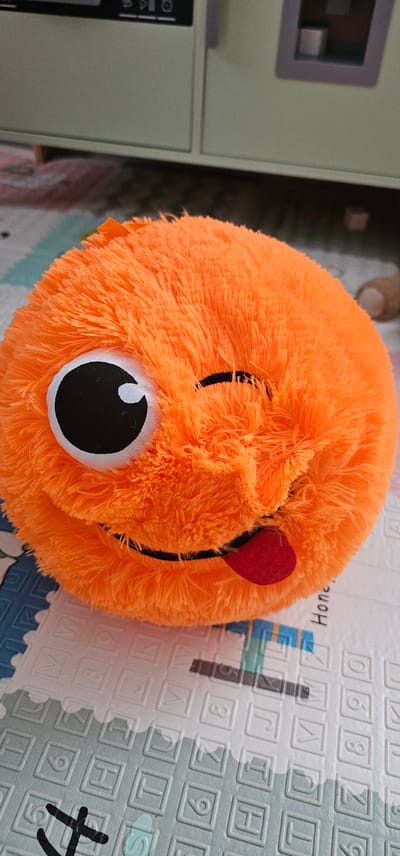 A close-up, eye-level, vertical shot captures an orange, fuzzy, spherical toy with a face. The toy has one large, open eye with a black pupil and a white highlight, and the other eye is closed in a wink. A black line forms a smile, and a red, felt tongue is sticking out. The toy rests on a white and blue patterned mat. In the background, a light green cabinet or shelf unit is visible. The location is not specified in the image.
