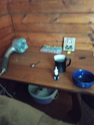 A blurred, grainy image shows a wooden table with various objects on it, set against a background of wooden walls and ceiling. In the upper left is a light blue object, possibly a hairdryer. To its right, a small framed picture of a person wearing a dark suit is leaning against the wall. In the center of the table, a black mug with white contents sits next to a small white dropper bottle with a black cap. To the right of the mug is a blue bowl. In the foreground, a purple bowl with handles is visible. The lighting is dim and the overall impression is of a cluttered, indoor space. The provided context indicates the location is Kirensk, Russia.