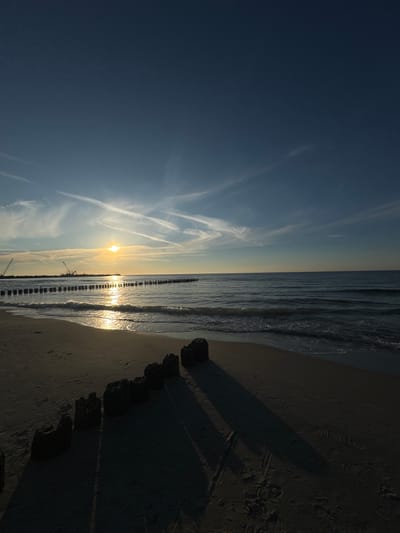 The image shows a sunset over the Baltic Sea at the beach in Ustka, Poland. The sun is low on the horizon, casting a golden glow on the water and the sky. Wispy clouds are scattered across the blue sky, illuminated by the setting sun. In the foreground, dark, weathered wooden posts emerge from the sand, creating a rhythmic pattern along the shoreline. The sand is wet from the receding tide, with footprints visible. In the distance, a line of similar wooden posts extends into the sea, forming a breakwater. Two construction cranes and some structures are visible on the far breakwater, suggesting ongoing development or maintenance. The waves gently lap at the shore, reflecting the warm light of the setting sun. The overall atmosphere is serene and peaceful, capturing the beauty of a coastal evening.