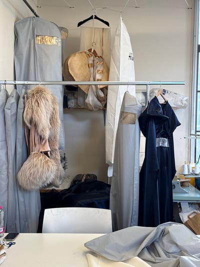 This image captures an indoor workshop or atelier in Arcueil, France, dedicated to garment production and storage. The space features light-colored walls and a window on the right providing ambient light. Two horizontal metal clothes rails are prominently displayed. The upper rail supports a grey garment bag with a clear window, a white garment bag partially marked with "A.B.", and a large, straw-colored circular textile, possibly a wide-brimmed hat, alongside other draped fabrics.
The lower, chrome-finish rail holds several additional grey garment bags. A voluminous, light brown/tan furry coat with a matching furry accessory hangs on the left side of this rail. To the right, a dark, possibly black, long garment resembling a costume or formal wear is displayed on a wooden hanger, characterized by a hooded design and a shimmering silver sequined band around the waist.
In the background, a light-colored shelf stores various items, and an industrial light blue sewing machine is visible on a table to the right, accompanied by several white thread spools. The foreground features a large white work table with a white chair pulled up to it. On the table's left, a partially consumed plastic water bottle, a dark smartphone, blue scissors, and a measuring tape are arranged. The right side of the table is covered with draped pieces of light grey and white fabric, indicating ongoing textile work. No individuals are present in the scene.