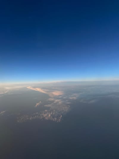 The image captures a view from an airplane at sunset, looking out at the horizon. The left side of the frame is dominated by a deep blue sky, gradually transitioning to a lighter shade closer to the horizon. On the right, the Earth's surface is visible, shrouded in atmospheric haze and clouds. The clouds are illuminated by the setting sun, casting warm, reddish-pink hues on their undersides, contrasting with the darker, shadowed portions of the landscape. The scene is characterized by a sense of vastness and tranquility, with the Earth appearing as a distant, dimly lit expanse beneath a vast sky. The time of day is clearly indicated by the sunset colors and the gradient of light in the sky. The location is Kansk, Russia. There is no visible text in the image.