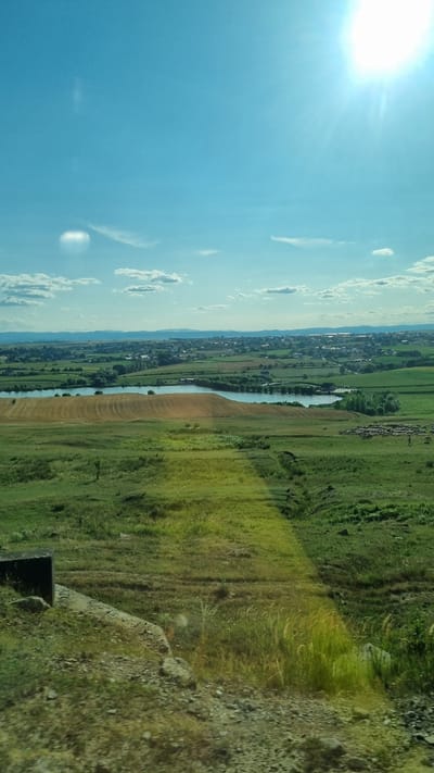 The image captures a panoramic view of a rural landscape under a bright, sunny sky. In the foreground, a rough, textured ground with scattered rocks and sparse vegetation leads to a gently sloping hill covered in green grass and some patches of dry, golden grass. A dark, rectangular concrete structure is visible on the left side of the foreground.

Further into the scene, a vast expanse of rolling green fields dominates the midground. A body of water, likely a lake or large pond, reflects the sky and is bordered by a field with distinct curved furrows, suggesting agricultural activity. Beyond the water, a flock of sheep is gathered in a field, with a few figures walking amongst them, indicating human presence and animal husbandry.

In the background, a town or village is nestled among the fields, characterized by a dense collection of buildings with various rooflines. The horizon is marked by a range of distant mountains, silhouetted against the pale blue sky. The sun is high in the sky, positioned in the upper right corner, casting a bright glare and visible rays, suggesting it is midday. The overall impression is one of a serene, expansive rural setting in the vicinity of Bulai, Romania.