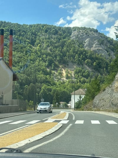 The image captures a view from inside a vehicle, looking forward through the windshield at a paved road. The road curves to the right and features a pedestrian crossing with white stripes and yellow painted islands. A silver car is visible on the road, approaching the pedestrian crossing. The surrounding environment is mountainous and lush, with dense green trees covering the slopes of a large hill that dominates the background. A rocky cliff face is also visible on the right side of the hill. On the left side of the road, there is a light-colored building with a dark-roofed section and two prominent reddish-orange chimneys with black tops. Further in the distance, another white building with a tiled roof can be seen nestled amongst the trees on the mountainside. The sky is blue with scattered white clouds. The image appears to be taken on a clear, sunny day. The scene is consistent with a road passing through a scenic, possibly rural or suburban area in a mountainous region. Given the context of Hauts de Bienne, France, this location is likely a road leading into or out of the town, showcasing its natural surroundings.