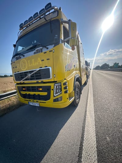 A yellow Volvo semi-truck, with the logo "ROLLA" prominently displayed on its front, is parked on the shoulder of a highway. The truck has multiple auxiliary lights mounted on its roof. The license plate reads "CT 9333 TH". The scene is bathed in bright sunlight, creating lens flare and casting a long shadow from the truck onto the asphalt. To the left of the truck, a field of dry wheat or similar crop is visible, bordered by a guardrail. The highway stretches into the distance, with a few other vehicles present. The sky is clear blue with a few wispy clouds. The location is identified as Trilistnik, Bulgaria.