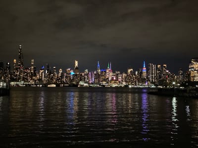 The image presents a panoramic nighttime view of the New York City skyline, observed from the waterfront in Weehawken, New Jersey, looking east across the Hudson River. The sky, occupying the upper third of the frame, is dark and overcast, showing no celestial bodies.
The central feature is the illuminated Manhattan cityscape. Numerous skyscrapers of varying heights and architectural designs are densely packed along the horizon. Prominent structures include the Empire State Building, identifiable by its top illuminated in red, white, and blue. To its left, another tall building exhibits a green and white illuminated pinnacle. The majority of buildings display warm yellow or orange interior lighting from their windows, while several feature additional architectural lighting in hues of blue, purple, and white, creating a vibrant urban glow.
The lower two-thirds of the image consist of the dark, rippled surface of the Hudson River. The water acts as a reflective medium, mirroring the city lights as elongated, shimmering vertical streaks of color—yellow, orange, blue, red, and purple—which distort slightly due to surface movement.
In the far left foreground, a dark, indistinct structure, consistent with a pier or a portion of a ferry terminal, is partially visible. On the far right foreground, a darker landmass or constructed barrier is present, marked by a single, illuminated light pole that casts a localized beam onto the water immediately adjacent to the shoreline. No individuals, overt actions, or moving vehicles are discernable within the depicted scene.