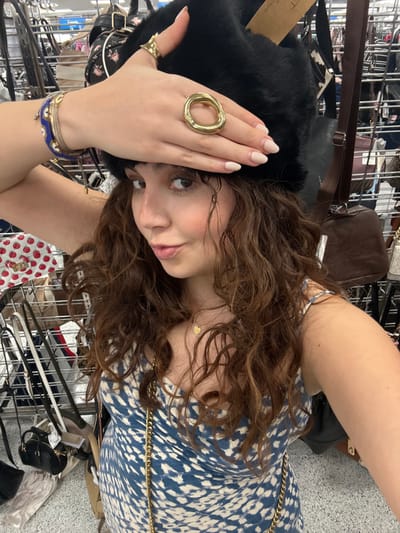 A woman wearing a blue patterned dress and a large black fur hat poses for a selfie indoors. She is wearing several rings, including one large gold ring, and multiple bracelets. She has brown curly hair and fair skin. A gold chain is draped across her dress. The background shows racks of purses and other accessories, suggesting a retail or thrift store environment.